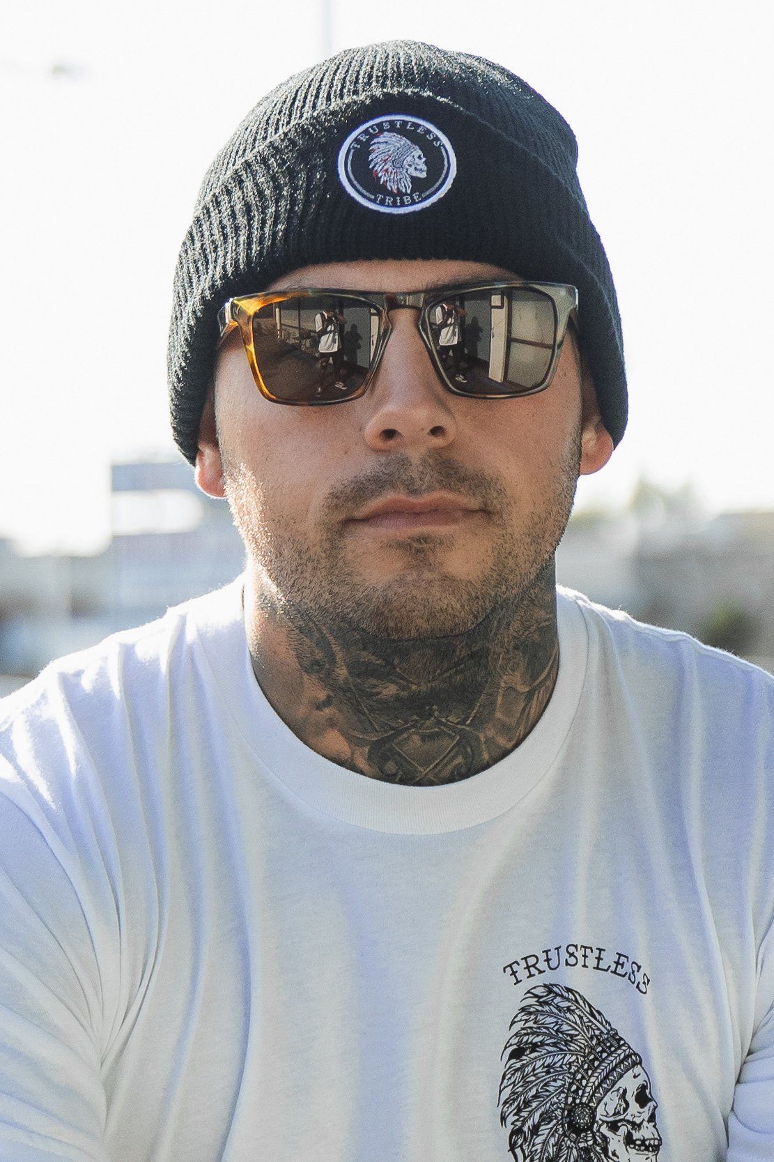 A man wears a Trustless Tribe acrylic beanie by Relentless Betrayal, sunglasses, and a white skull TRUSTLESS shirt. Tattoos coil up his neck as he stands in a sunny outdoor scene—a modern anarchist look.