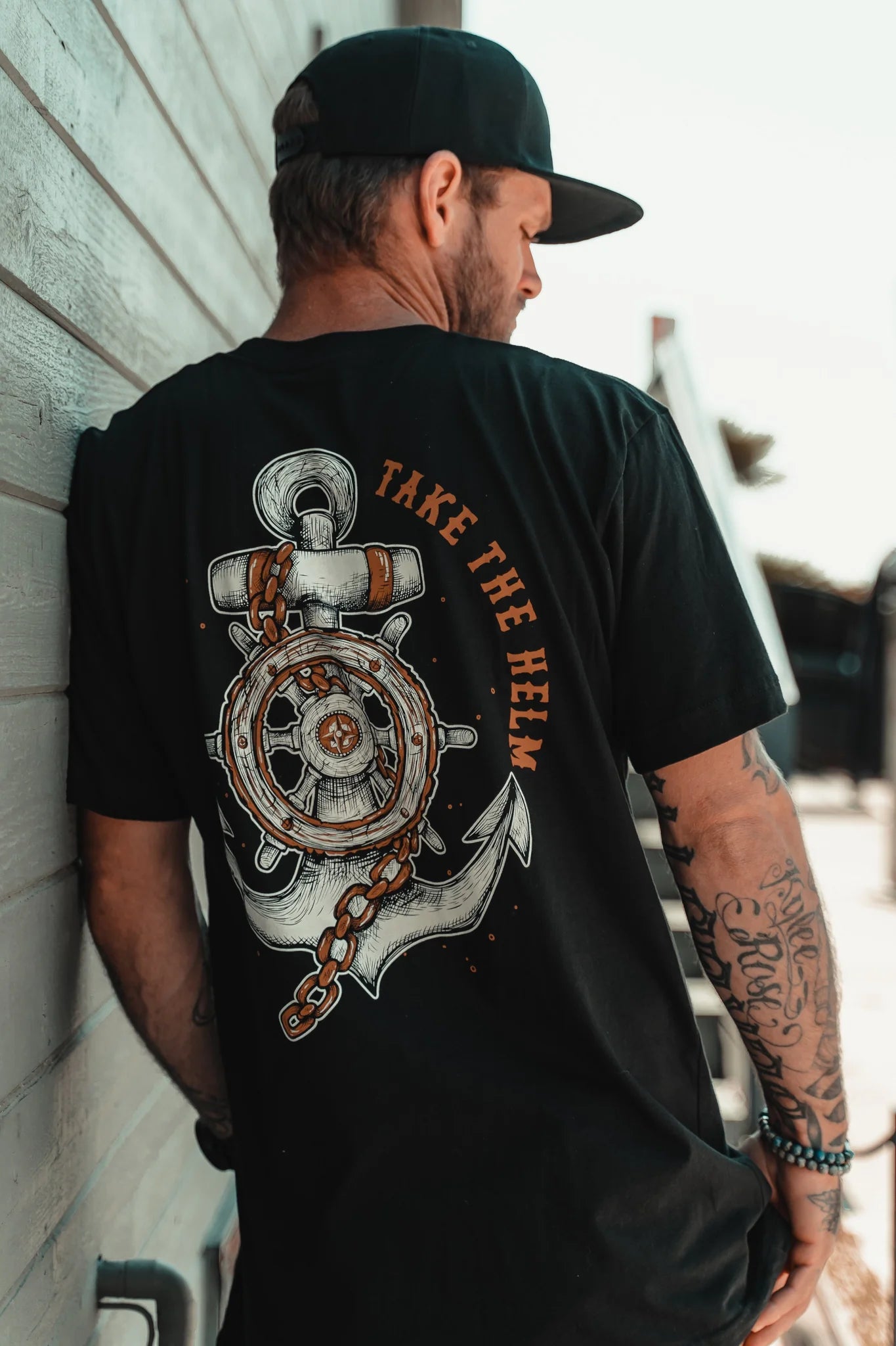 A tattooed man stands facing a wall, wearing Relentless Betrayals classic black Take The Helm Tee with a prominent anchor and steering wheel design on the back, adorned with TAKE THE HELM. He complements the look with a black cap against a blurred background, highlighting this unisex T-shirt style.