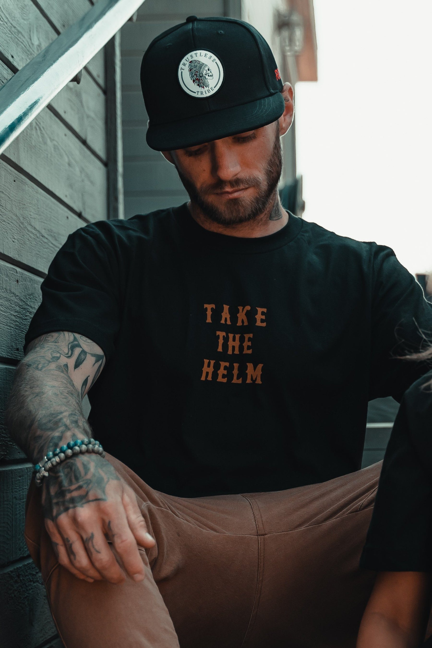 A tattooed man in a black cap and the Relentless Betrayal Take The Helm Tee sits outdoors by a wooden wall, looking down with his arms resting on his knees.