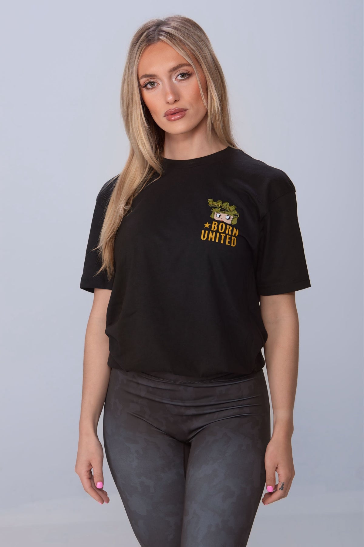A person with long blonde hair stands against a neutral background, wearing a black Born United Private Label t-shirt and Midnight Camo Leggings. Their hands are relaxed by their sides as they look directly at the camera.