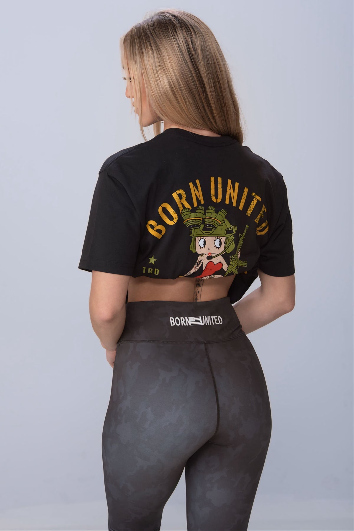A woman with long hair, wearing a black crop top with a cartoon graphic and BORN UNITED in bold yellow, pairs it with high-waisted Midnight Camo Leggings from Born United Private Label, showcasing a sleek black camo design.