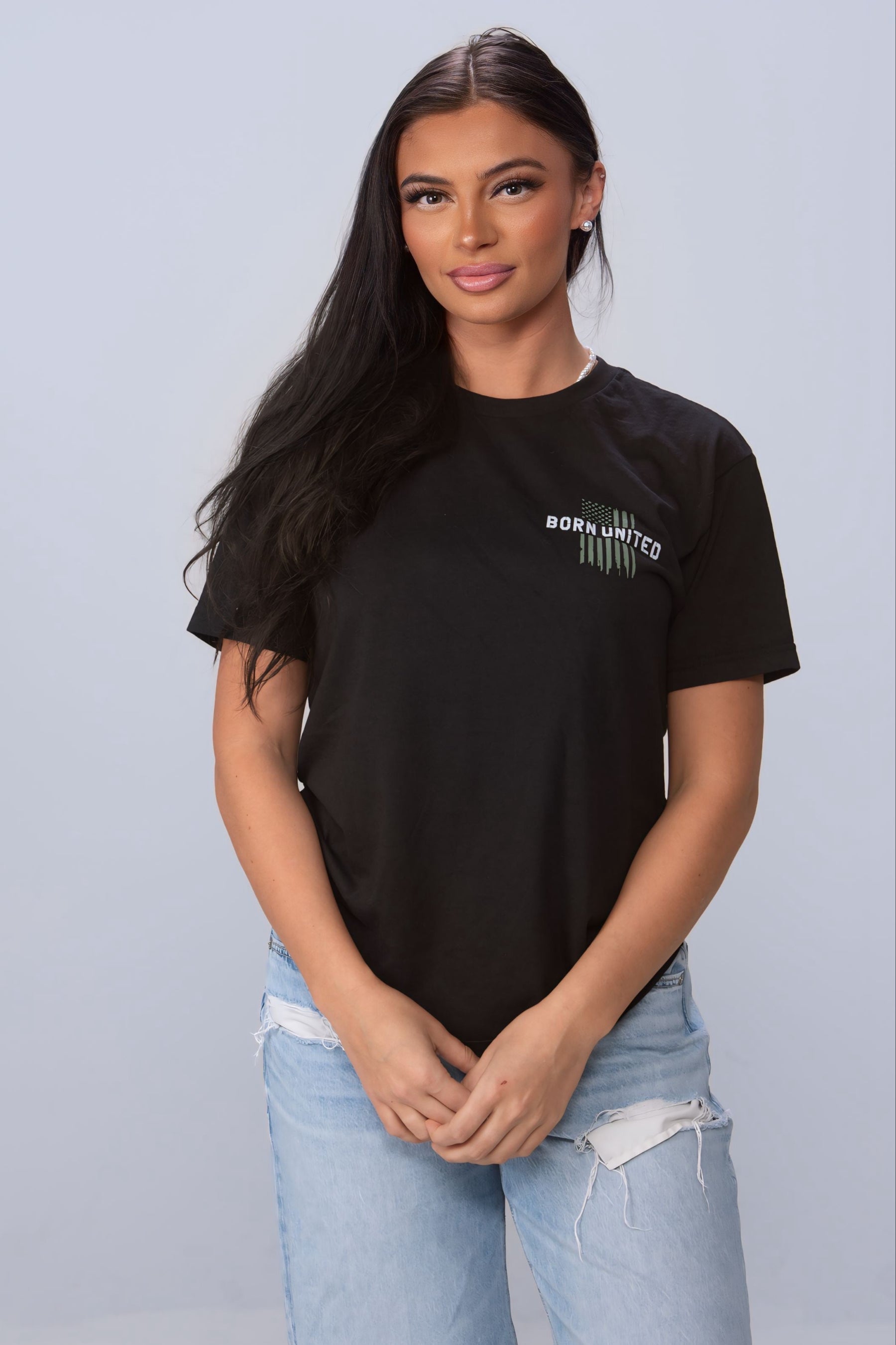 A person with long dark hair stands against a plain background, wearing light blue distressed jeans and a black American Heroes Tee by Born United Private Label, their hands clasped as a silent tribute to American heroes and first responders.