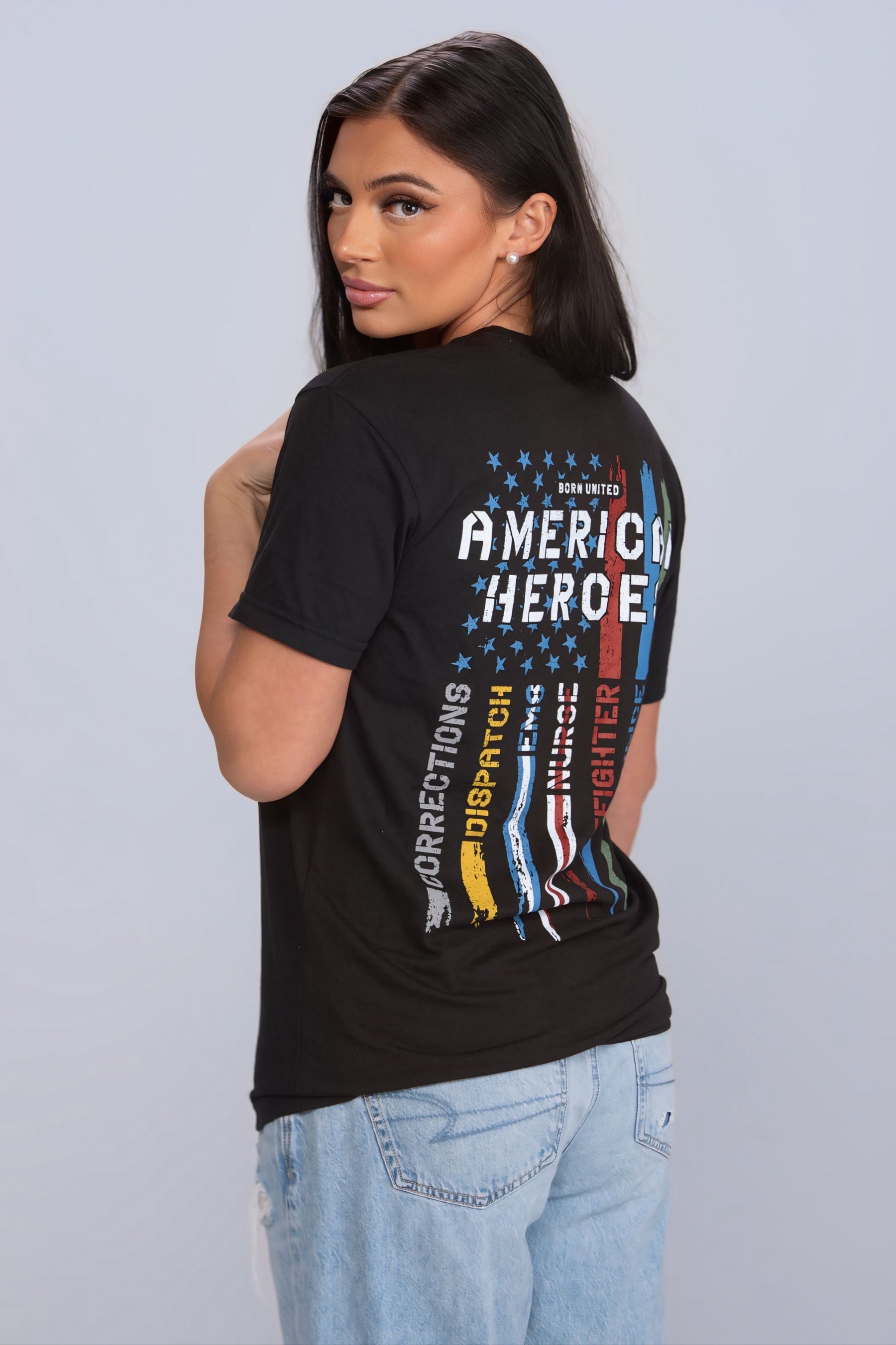 A woman wears the black Born United Private Label American Heroes Tee, featuring colorful text saluting jobs like military and firefighter on the back, paired with light blue jeans, as she glances over her shoulder against a light gray background.