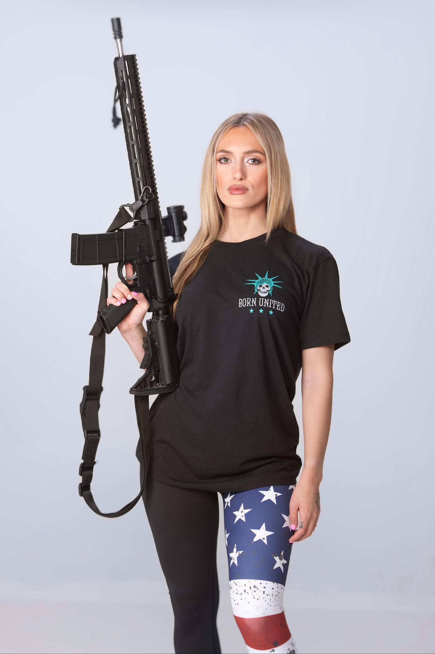 A person with long blonde hair, dressed in a black Liberty or Death tee by Born United Private Label and leggings with an American flag design, holds a rifle. The simple backdrop accentuates their embodiment of freedom and patriotism.