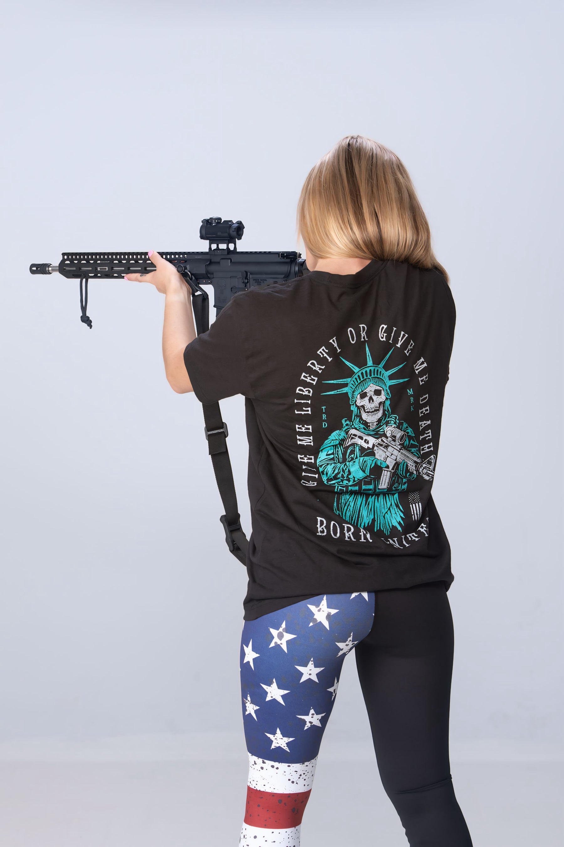 A long-haired individual faces away with a rifle, wearing Born United Private Labels Liberty or Death Tee and star-spangled leggings against a light gray backdrop, embodying the spirit of liberty.