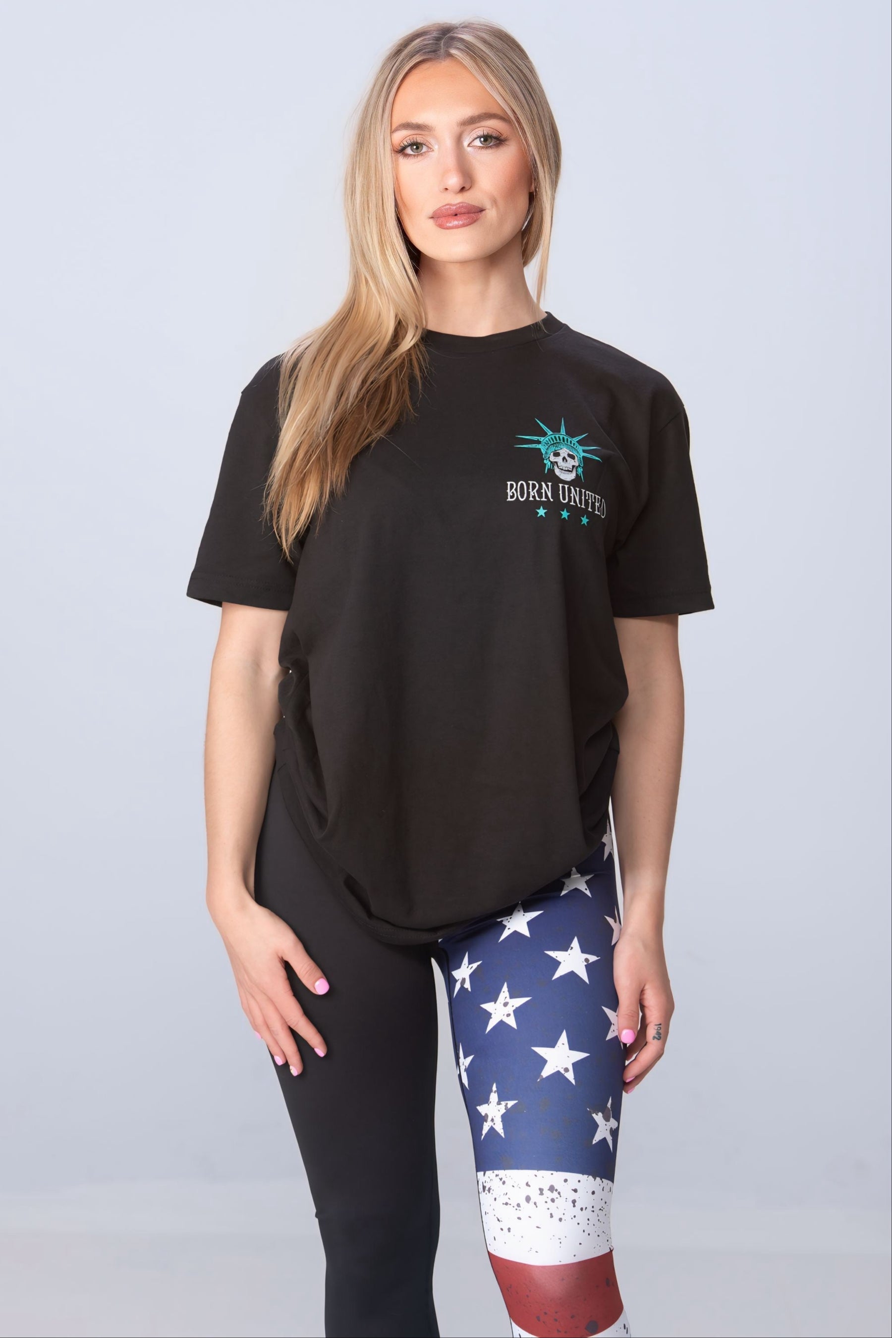 A woman stands against a light gray backdrop, wearing a black Born United Private Label t-shirt and American Savage Leggings featuring an American flag design on one leg. Her long blonde hair cascades down, enhancing her confident and stylish look.