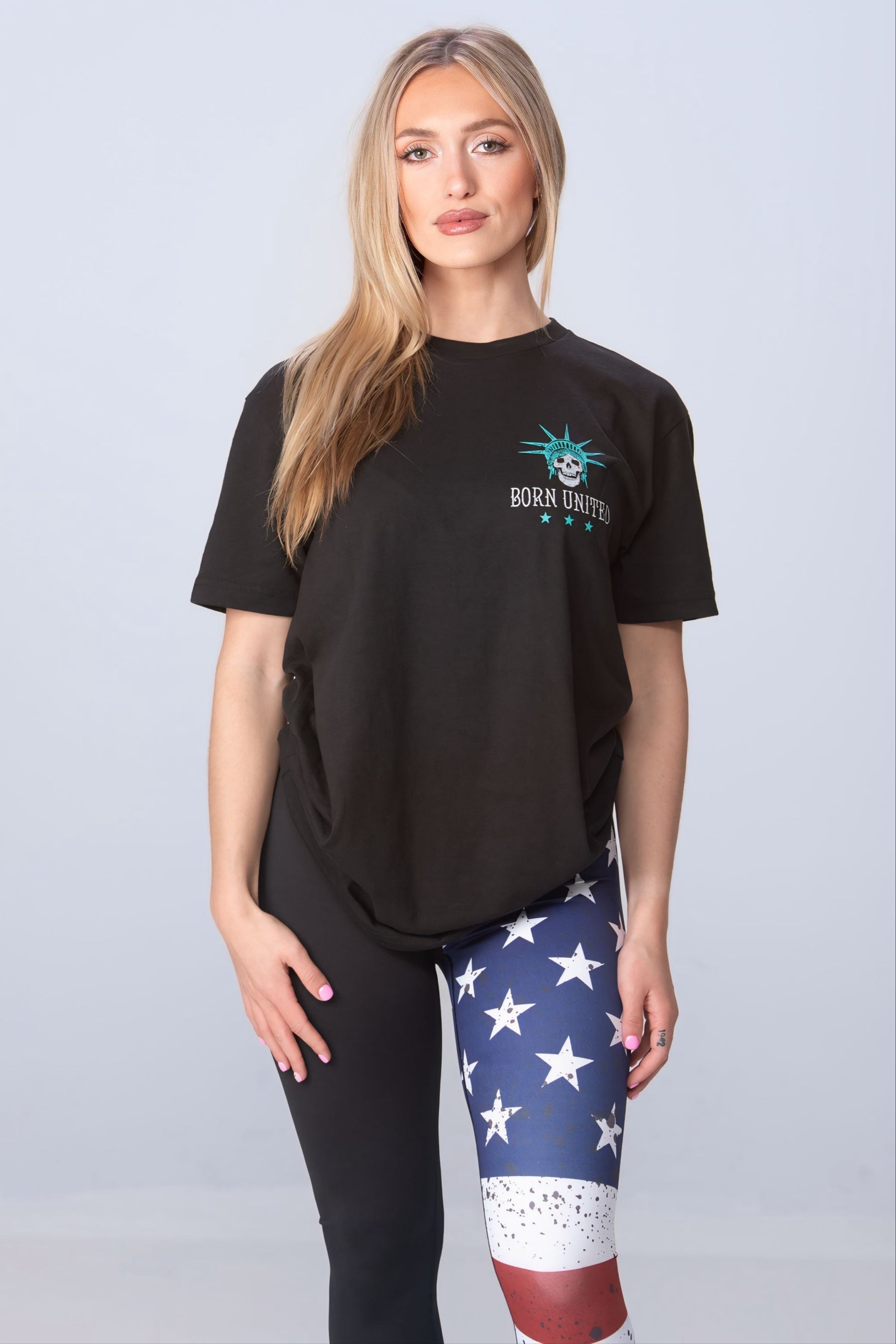 A long-haired blonde wears a black Liberty or Death T-shirt by Born United and leggings with an American flag on one leg, capturing the essence of freedom against a simple grey backdrop.