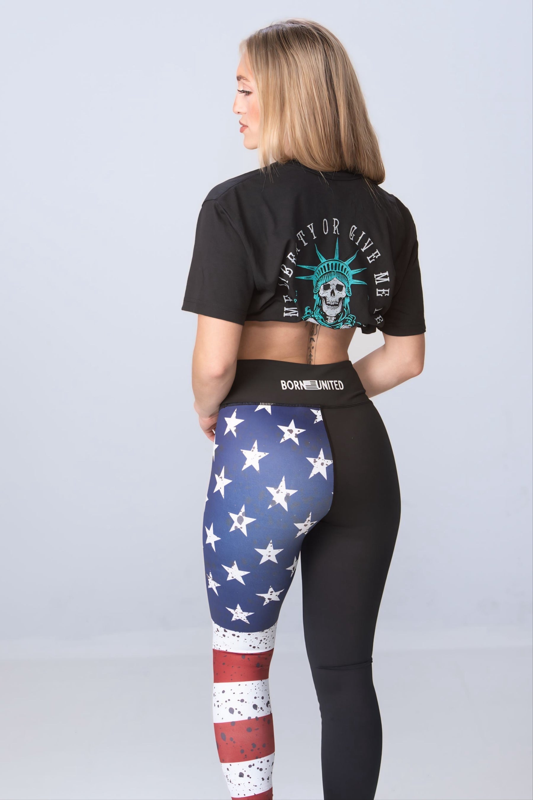 A woman with long blonde hair wears a Born United Private Label Liberty or Death Tee, black and cropped with a back graphic. She pairs it with two-tone leggings; one leg with a stars and stripes freedom pattern, the other solid black. The backdrop is plain light gray.