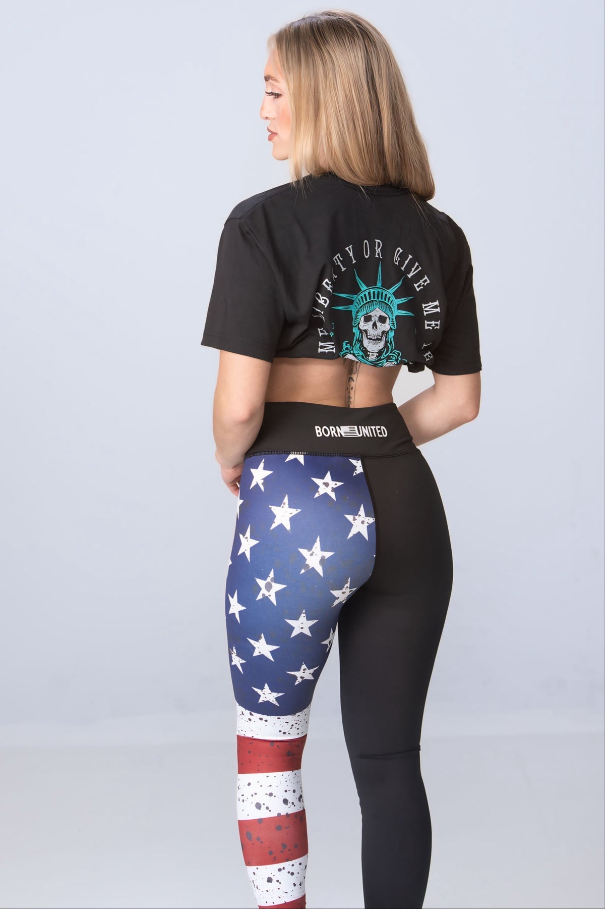 A long-haired blonde woman models eye-catching American Savage Leggings by Born United Private Label, featuring one leg with an American flag design and the other solid black. She pairs them with a black graphic crop top against a plain light background.