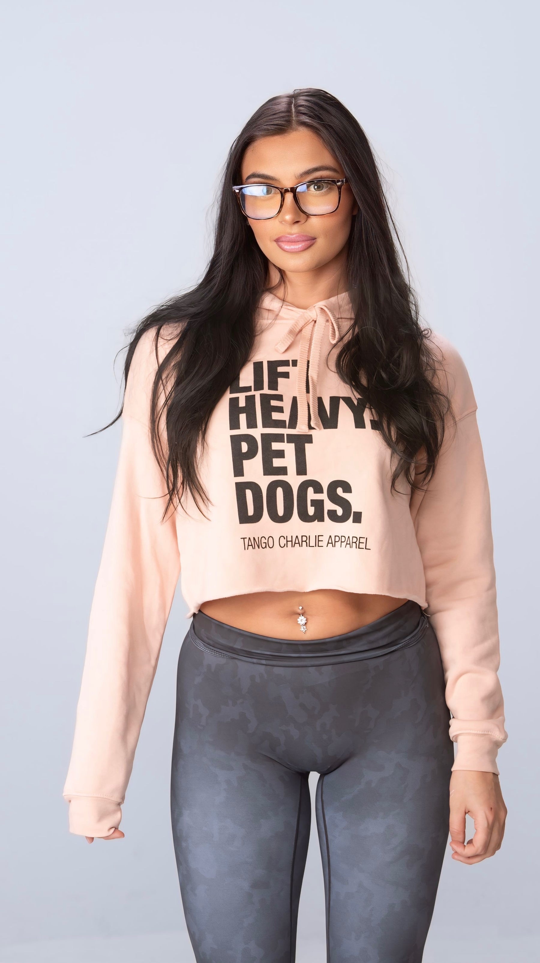 A woman with long dark hair and glasses wears a peach Lift Heavy, Pet Dogs crop hoodie from Tango Charlie Apparel paired with gray camo leggings. Set against a simple backdrop, she exudes relaxed style and comfort effortlessly.