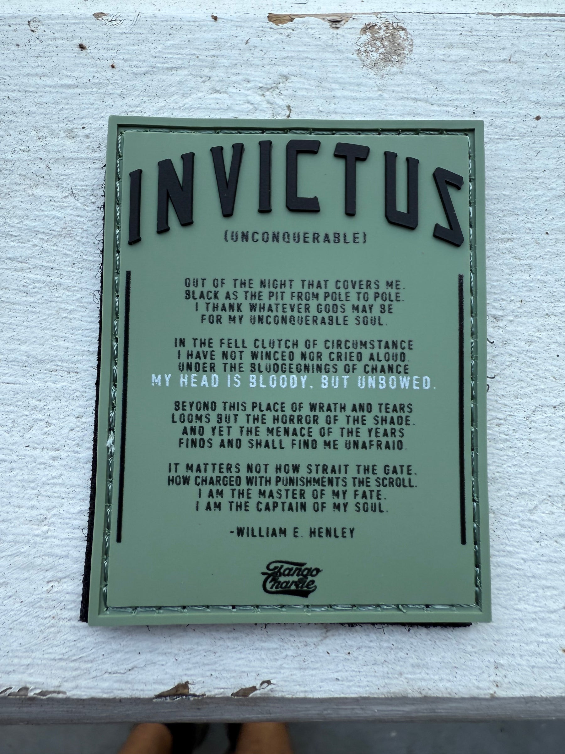 An image features the Invictus PVC Patch by Tango Charlie Apparel on a white wooden surface, displaying William E. Henleys poem Invictus. The title Invictus (Unconquerable) is prominently in large letters at the top, with feet partially visible at the bottom.