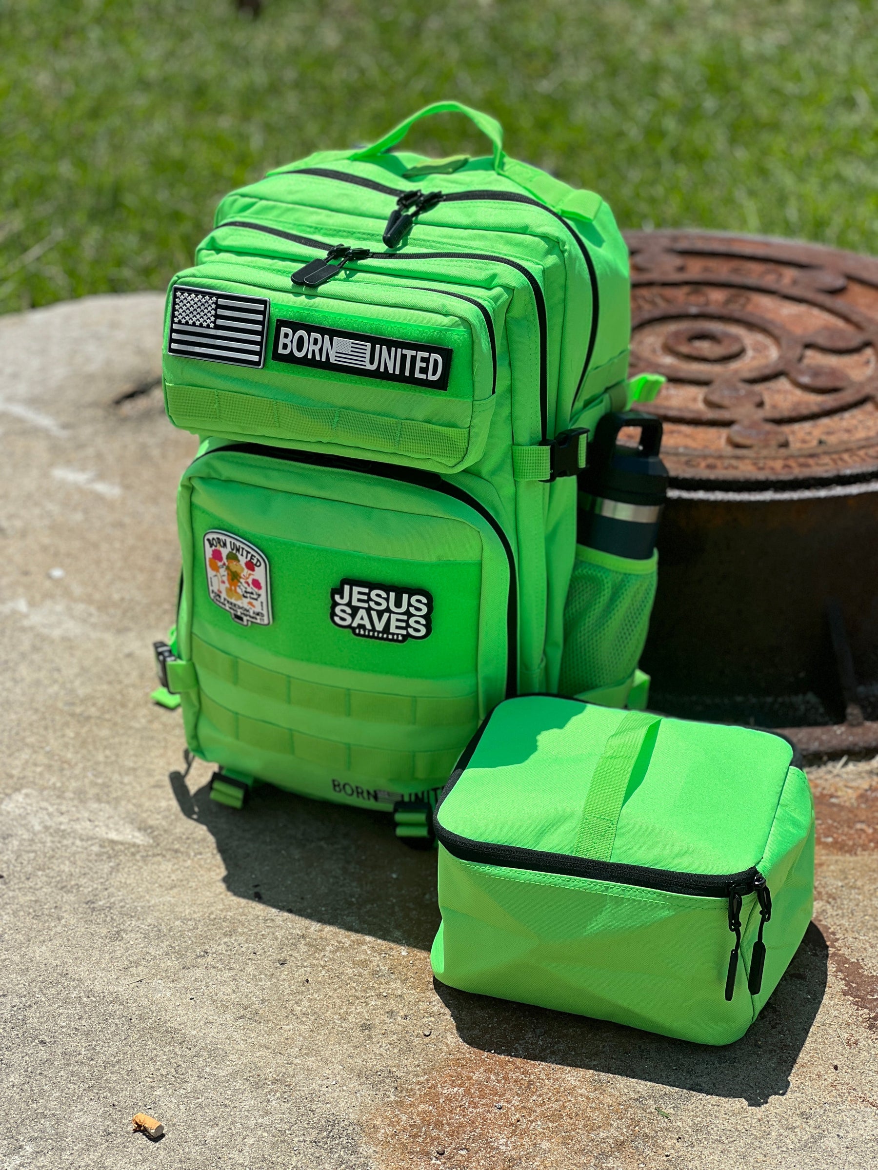 The Key Lime Tac-Pac - 45L by Born United Private Label, featuring BORN UNITED and JESUS SAVES patches, a side water bottle, and matching lunch bag, sits on concrete beside a rusty cover with grass in the background.