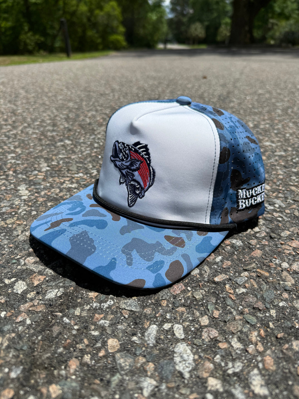 A Monster Bass Merica Water Camo Bucket by My Store, featuring blue and white camo, a fish embroidery on the front, and Muck Buck on the side, rests on sunlit asphalt with blurred trees in the background.