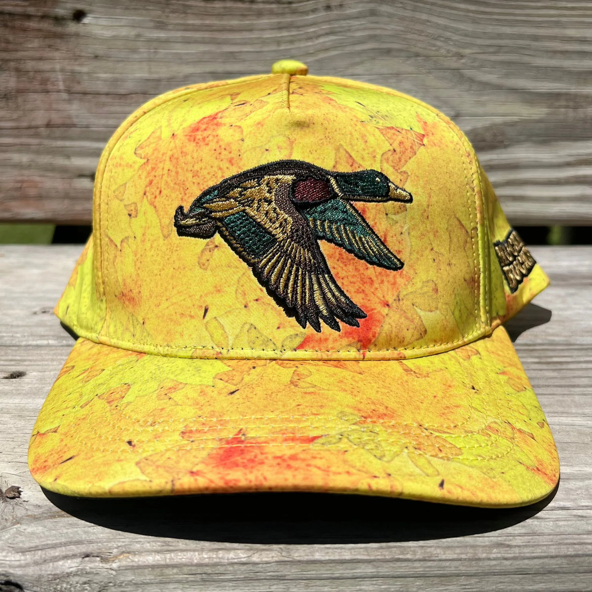 The Golden Leaf Camo Duck Bucket by Mucker Bucket exudes duck hunting charm, featuring a yellow camo leaf pattern and an embroidered duck in flight on the front. Its displayed on a wooden surface to highlight the standout design.