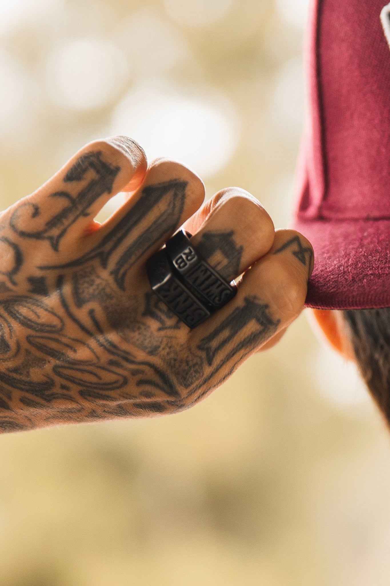 A tattooed hand adjusts a maroon cap, showcasing Relentless Betrayals Polished Black Sink or Swim Ring. With numerous tattoos adding artistic flair, the background softly blurs with earthy tones.