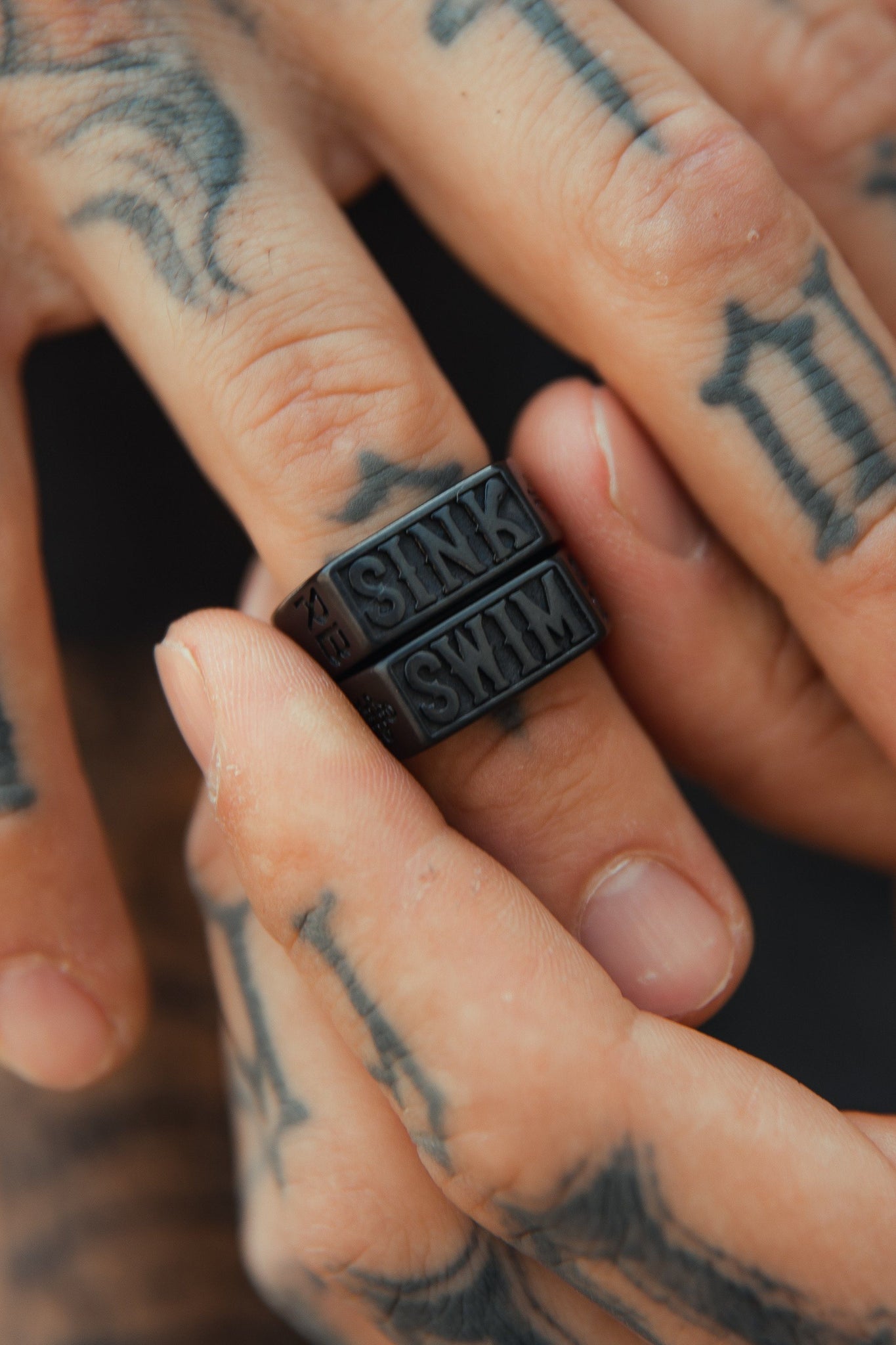 A close-up of tattooed hands shows two polished black stainless steel rings by Relentless Betrayal on the finger, with SINK on top and SWIM below. The bold tattoos provide a textured contrast to these ocean-themed Sink or Swim rings.