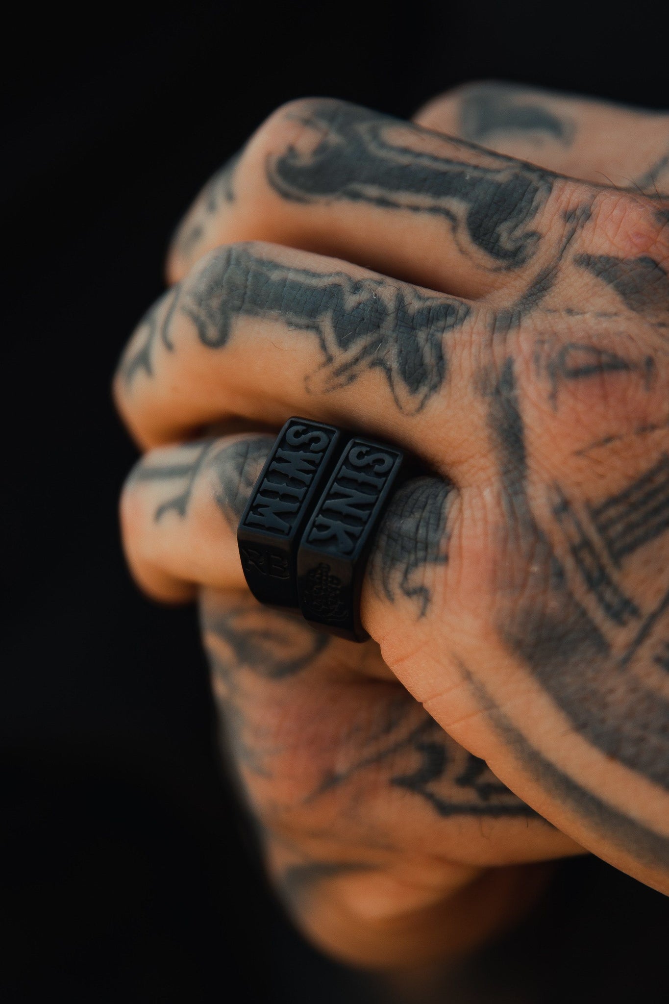 Close-up of a tattooed hand with white designs, featuring handcrafted Polished Black Sink or Swim Rings by Relentless Betrayal with SINK engraved on them. The dark background highlights intricate tattoos and ocean-themed jewelry details.