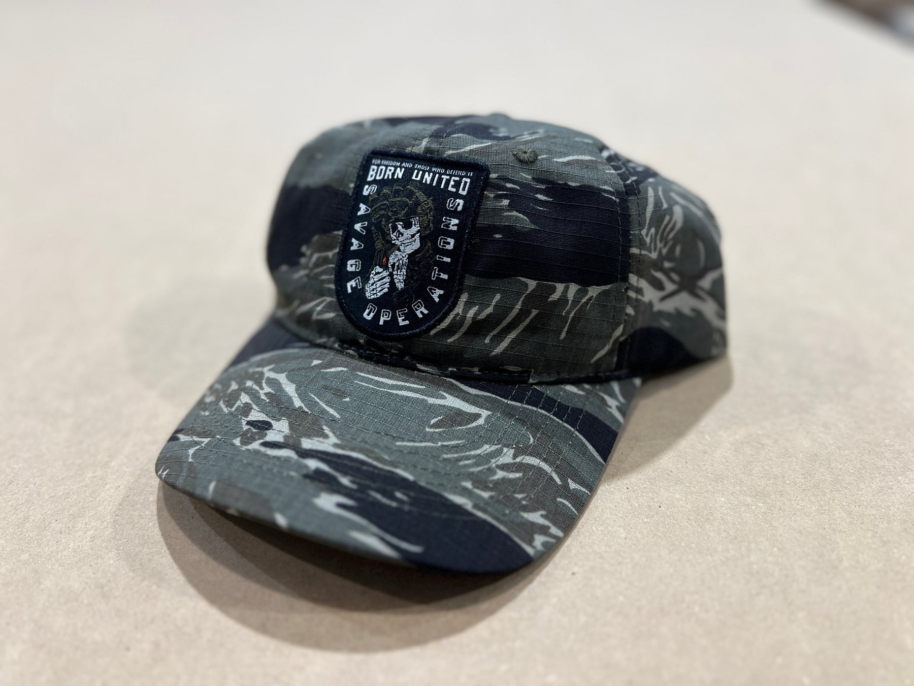 The Born United Private Label "Born United Buckle Hat - Savage Operations," a camo baseball cap featuring an eagle emblem and “BORN UNITED WAR OPERATIONS” patch, is displayed on a light-colored surface.