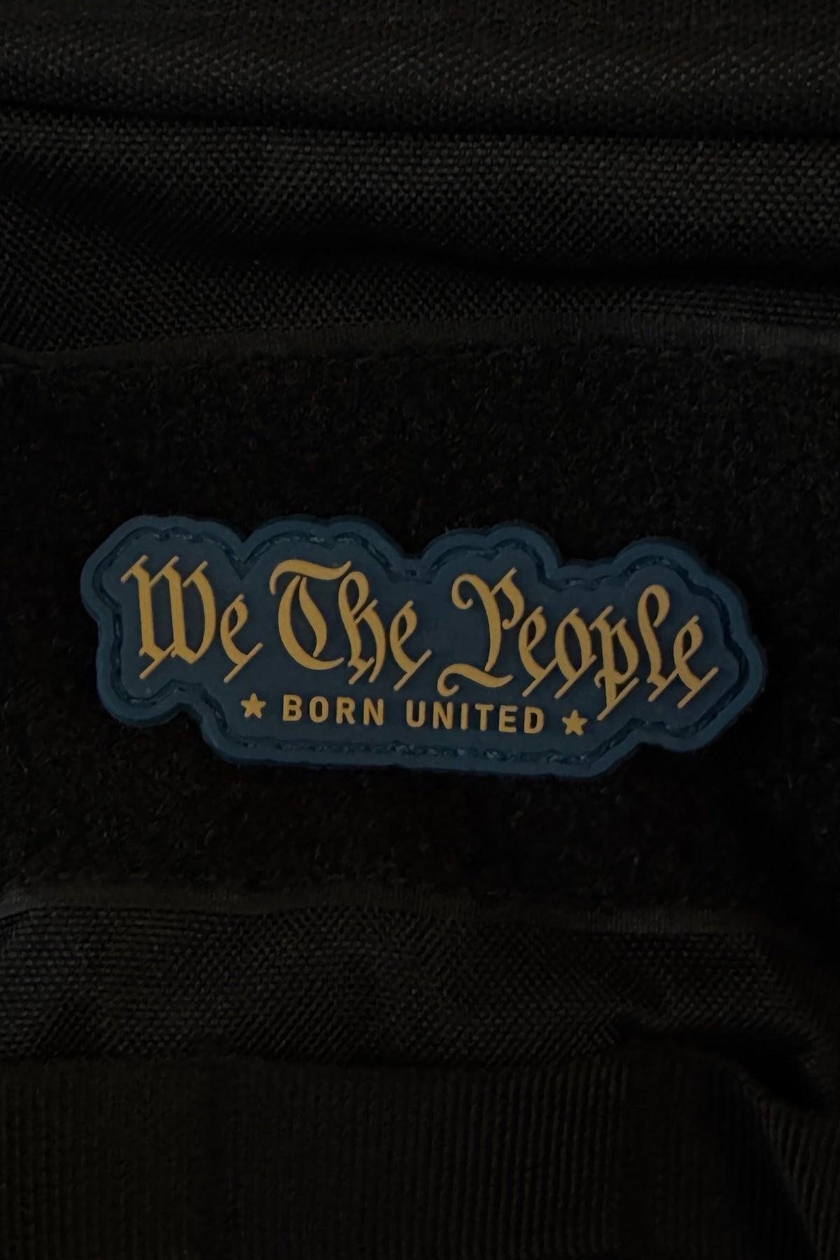 A bold 3-inch “We The People PVC Patch” by Born United Private Label features gold script reading “We The People BORN UNITED” on a dark, star-studded background, and attaches securely to black fabric with Velcro.