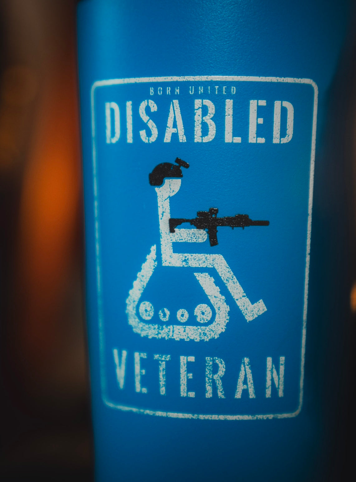 A blue sign features a wheelchair with tank tracks and a person holding a rifle, labeled “Born United Disabled Veteran”—a powerful design for the 20oz Disabled Veteran Tumbler from Born United Private Label.