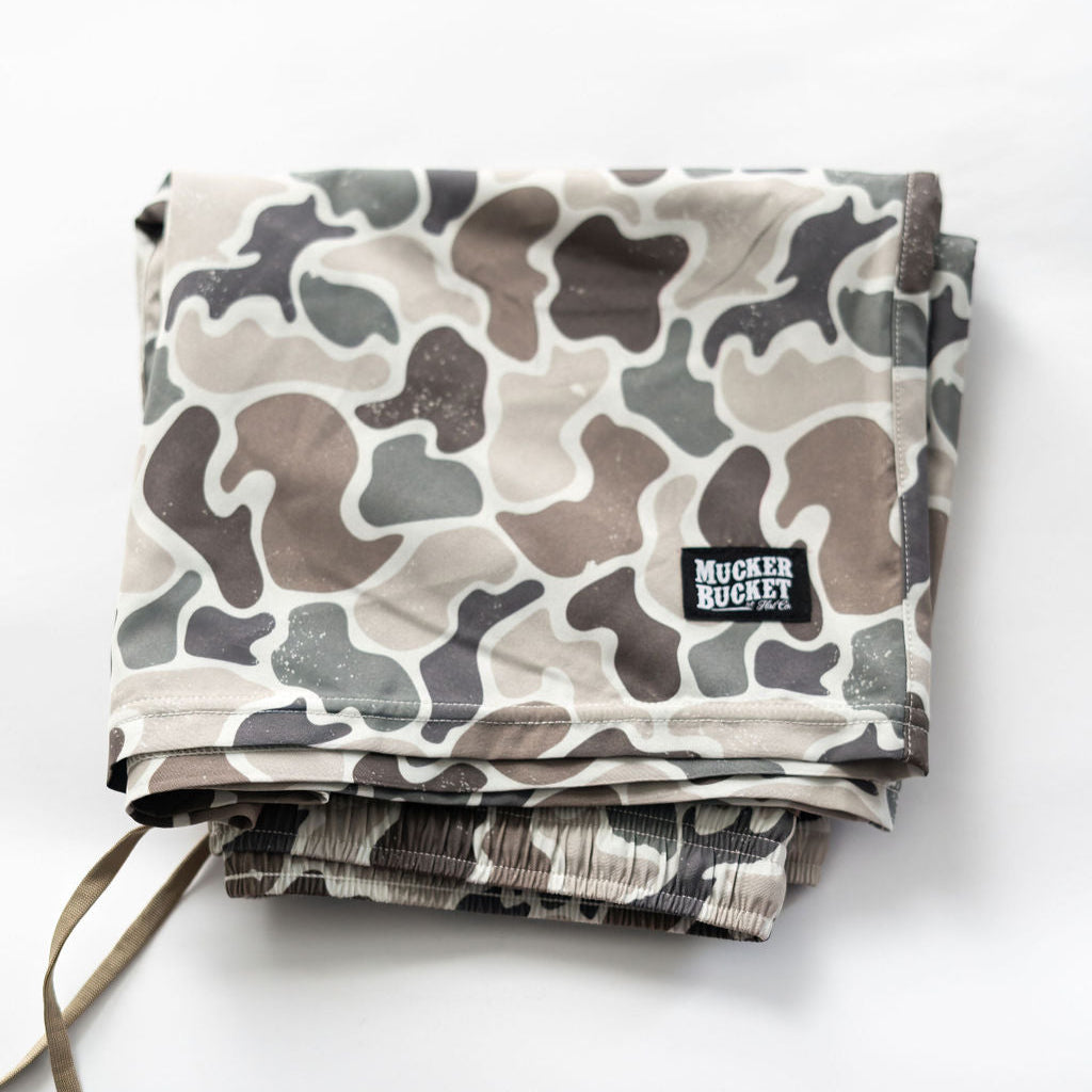 Folded gray and brown camouflage-pattern Mens Performance Athletic Shorts from Mucker Bucket are neatly arranged on a white surface, resembling a ducks gliding feathers. A small black patch reads Mucker Buckett.