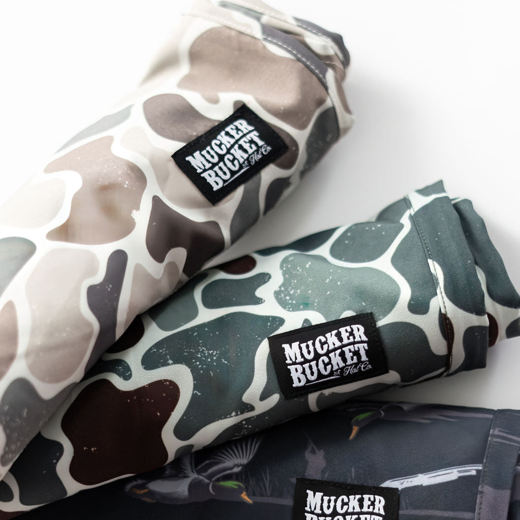 Three Mucker Bucket Mens Performance Athletic Shorts, Duck It collection, feature camo patterns in light beige, olive green, and dark green, stacked on a white surface—ideal for duck enthusiasts who love blending into nature.