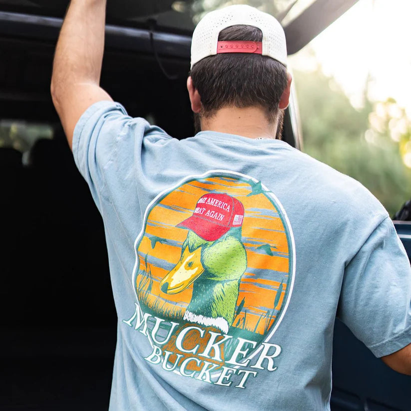 A man in a cap and light blue Smart Duck Tee by Mucker Bucket with a duck graphic wearing a red hat stands against blurred greenery and part of a car. The ring-spun cotton shirt offers a premium relaxed fit.