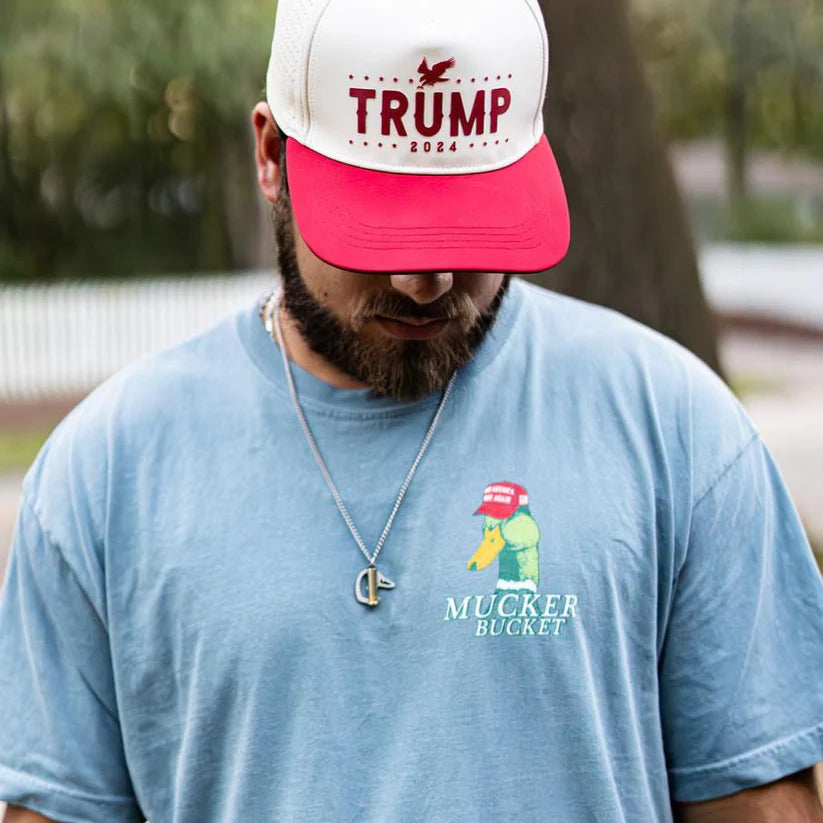 Wearing a light blue premium relaxed fit Smart Duck Tee by Mucker Bucket, featuring a cartoon duck with a red cap, a man glances down. His red and white cap reads Trump 2024, while a chain necklace rests on the garment-dyed 100% ring-spun cotton fabric.