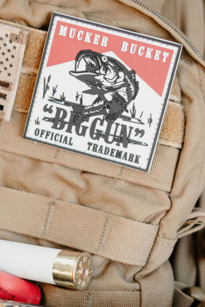 A tan tactical vest features the "Biggun" PVC Patch by Mucker Bucket, displaying a fish graphic and the text "Biggun Official Trademark." Visible Velcro patches include a shotgun shell and an American flag.