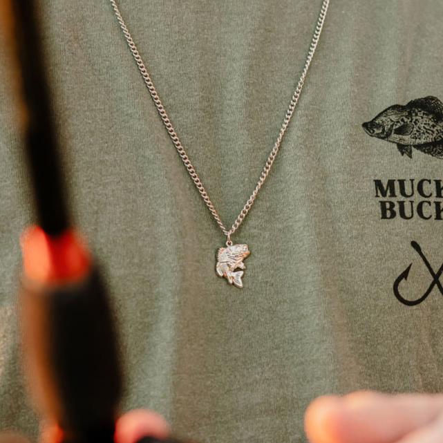 A close-up shows a person wearing Mucker Buckets Big Bass America Necklace, holding a fishing rod. Theyre in a green shirt with a fish illustration and the phrase MUCH BUCK. The necklace is the focus.