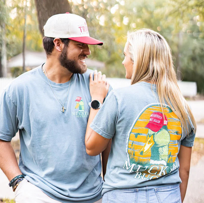 A smiling couple outdoors wears matching Mucker Bucket Smart Duck Tees, crafted from 100% ring-spun cotton with colorful back graphics. The man dons a red and white cap, while the woman displays her Apple Watch amid trees and a house in a sunny, relaxed setting.