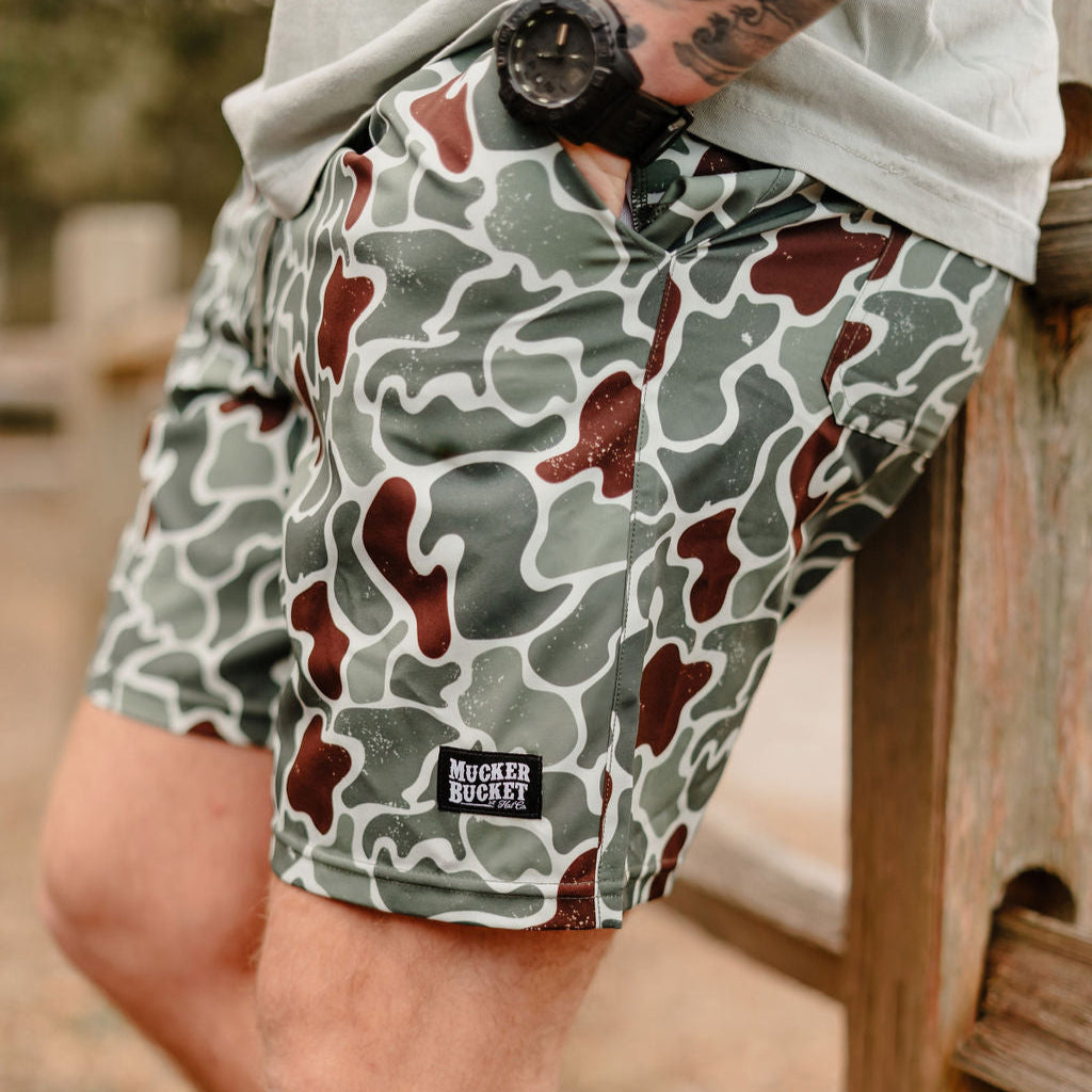 A person wearing Mucker Buckets Mens Performance Athletic Shorts, 5.5- Duck King in green and brown camo-pattern, a watch, and a light shirt is leaning on a wooden surface outdoors.