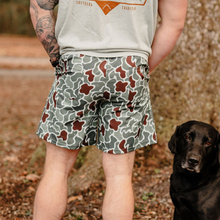 A person wearing a light gray shirt and Mucker Buckets Mens Performance Athletic Shorts in Duck King camo stands next to a black dog. The tattooed individual is outdoors by a tree, as the dog looks toward the camera.