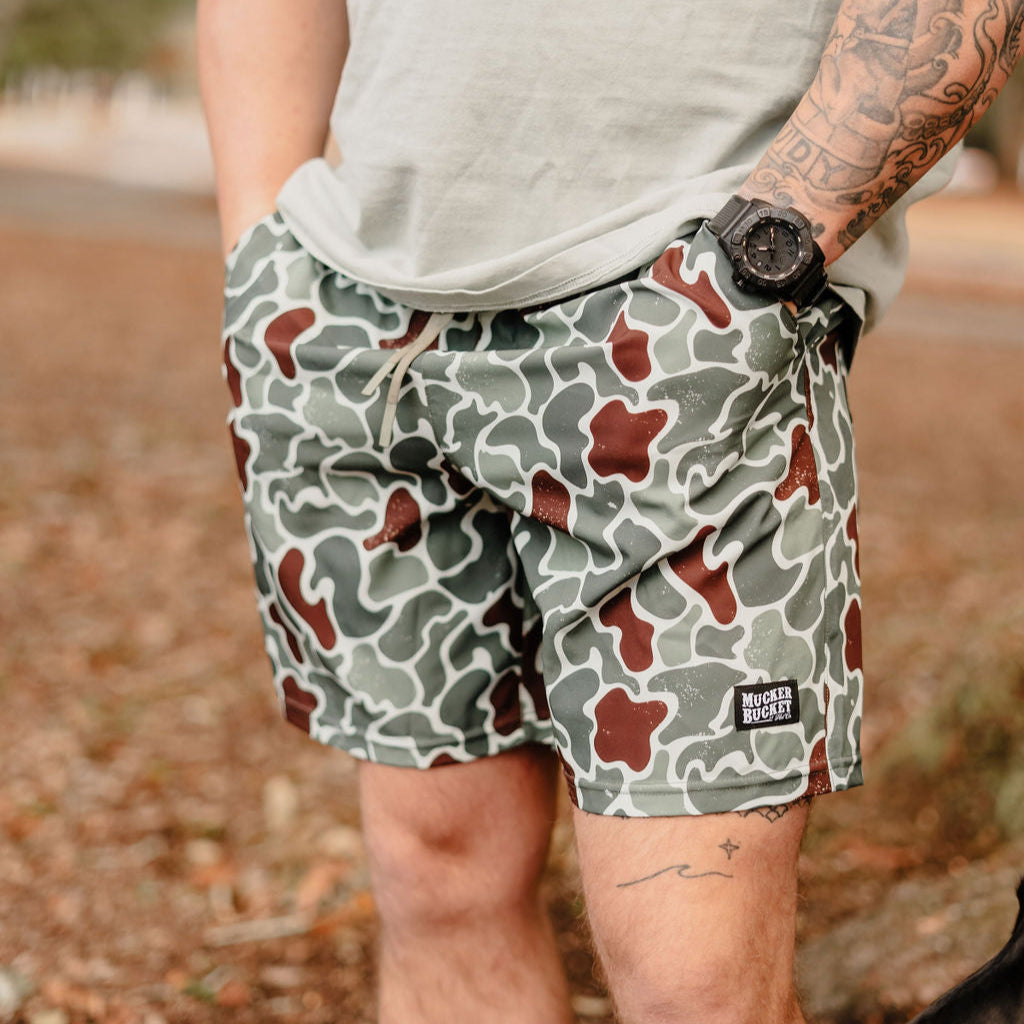 A person wearing Mens Performance Athletic Shorts | 5.5- Duck King by Mucker Bucket has hands in pockets, with a watch and tattoos visible on their left arm against a backdrop of blurred foliage and ground.