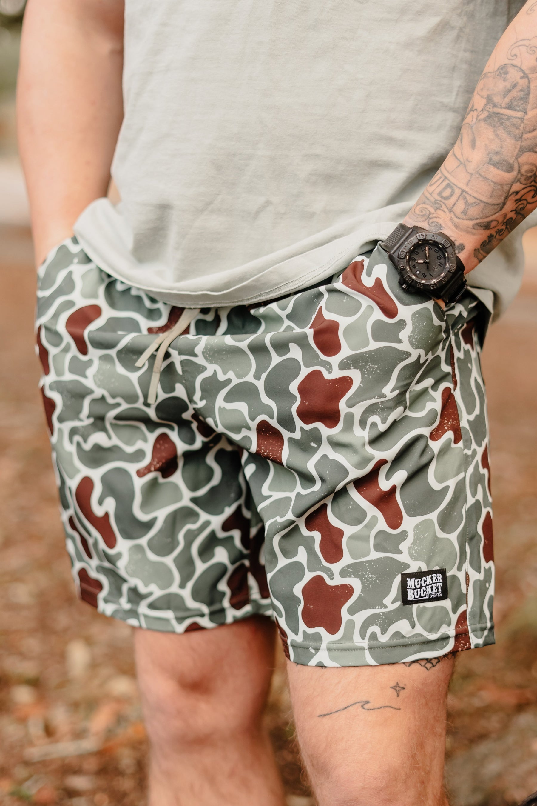 A person is wearing green and brown camouflage Mens Performance Athletic Shorts | 5.5 - Duck King by Mucker Bucket with hands in pockets, a light green shirt, a black watch on their left wrist, and visible tattoos on their left arm.