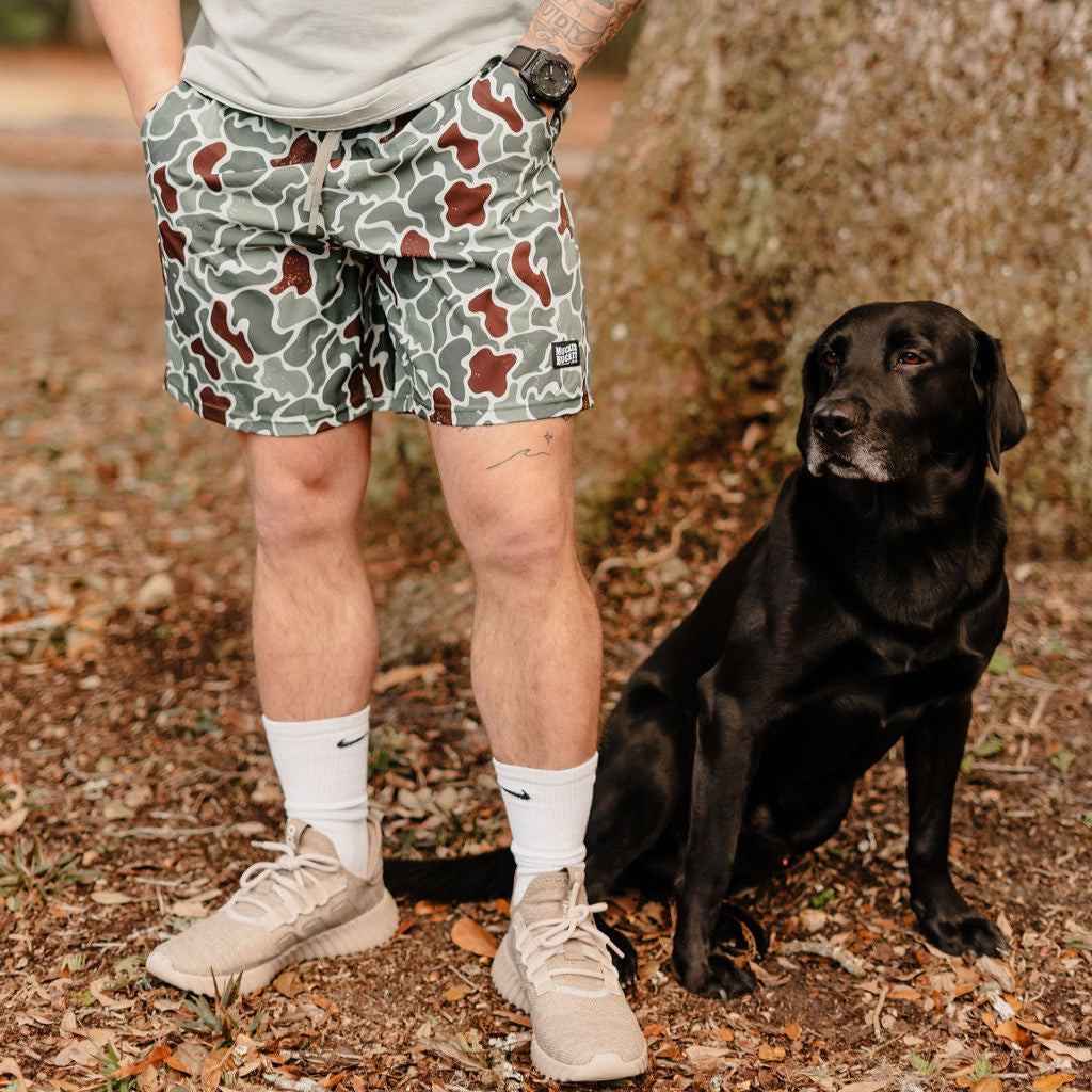 A person in Mucker Bucket Mens Performance Athletic Shorts | 5.5- Duck King stands beside a black Labrador on the ground, wearing beige sneakers and white socks, with trees and fallen leaves in the background.