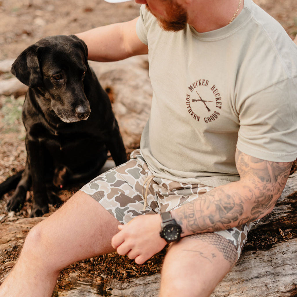 A tattooed man in a beige T-shirt and Mucker Buckets Duck It shorts sits on a log with his calm black Labrador in an outdoor setting, both seemingly savoring the peaceful moment.