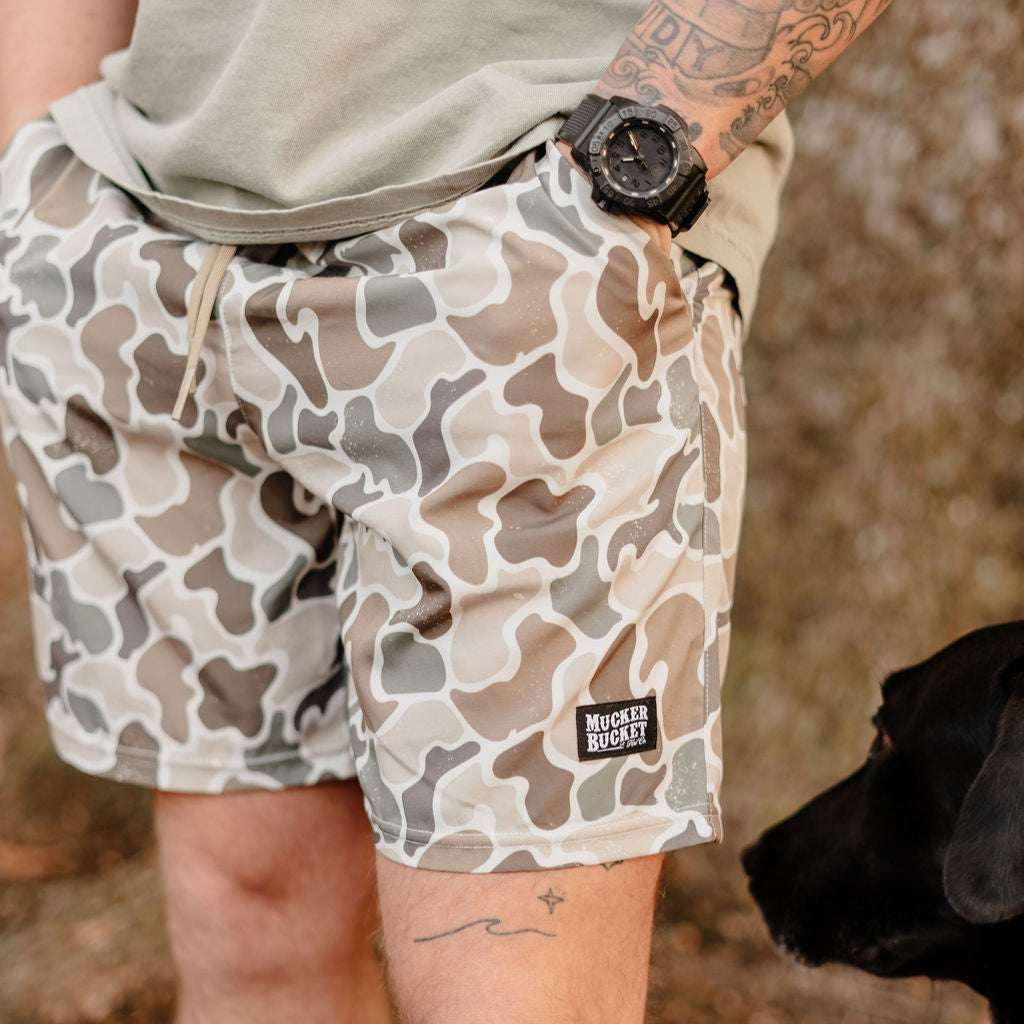 Wearing Mucker Buckets green Mens Performance Athletic Shorts in Duck It style and a light gray shirt, a person stands with their black dog. A tattoo decorates their left arm and a black watch sits on their wrist, perfectly blending practicality and style.
