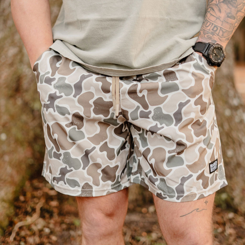 Donning Mens Performance Athletic Shorts by Mucker Bucket and a beige t-shirt, the person stands outdoors with a left-arm tattoo and black watch. Amid the peaceful treescape, they effortlessly blend into nature like a discreet duck.