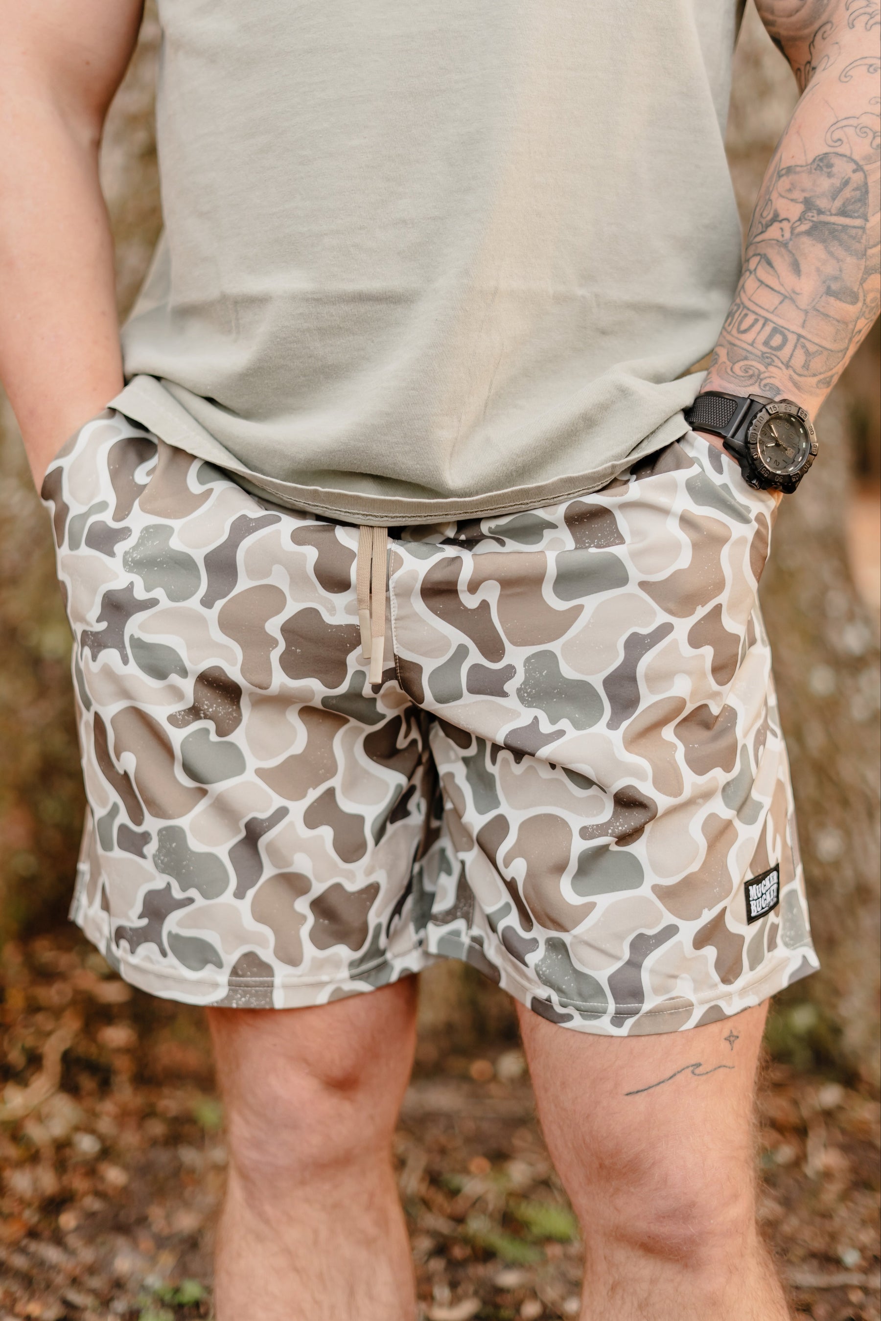 A person stands outdoors, hands in pockets, wearing a light green shirt and Mucker Buckets Mens Performance Athletic Shorts in the camouflage Duck It pattern, perfectly matching their tattoos and outdoorsy wristwatch. The blurred greenery behind them enhances the natural vibe.