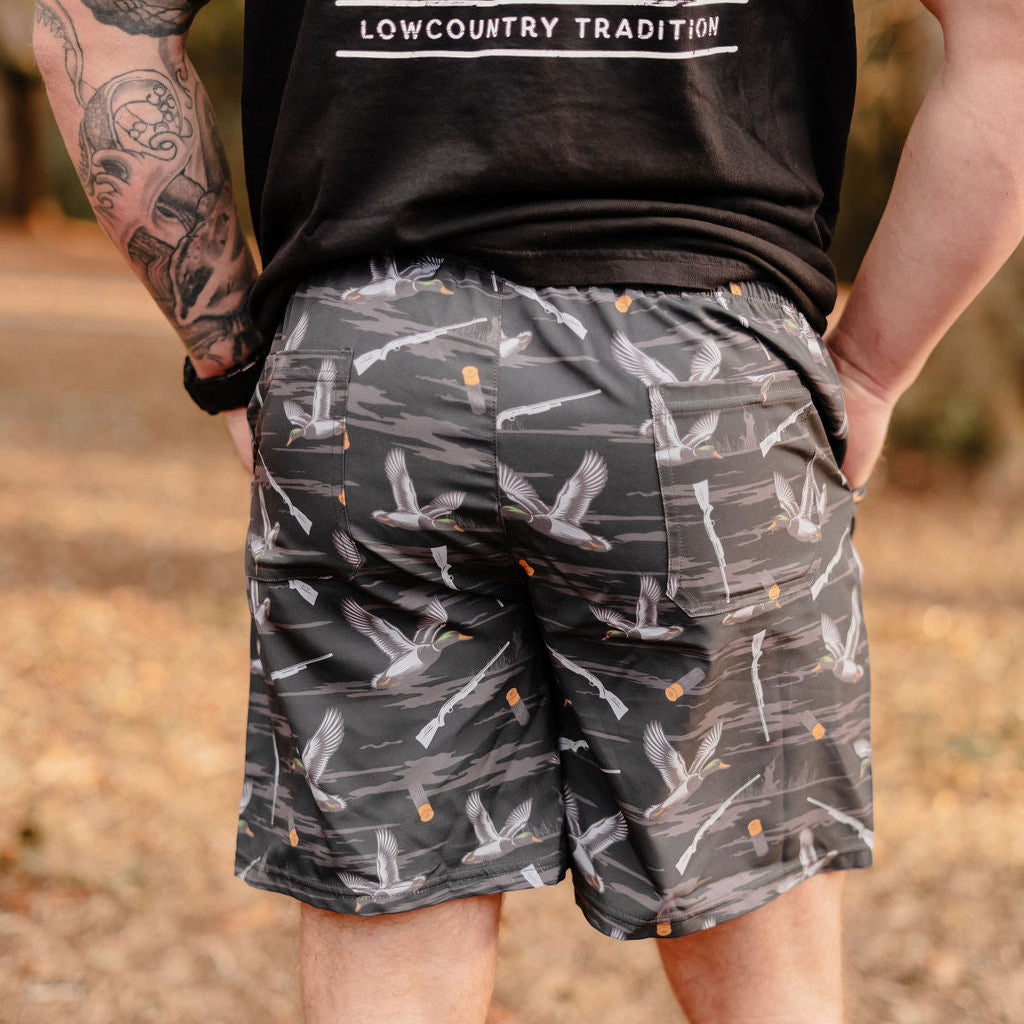 A person stands outdoors in a black t-shirt with LOWCOUNTRY TRADITION and Mucker Buckets Mens Performance Athletic Shorts | 5.5- Duck, Duck Boom, featuring flying bird designs and orange dots. They have tattoos on their left arm and hands in pockets against a blurred natural background.