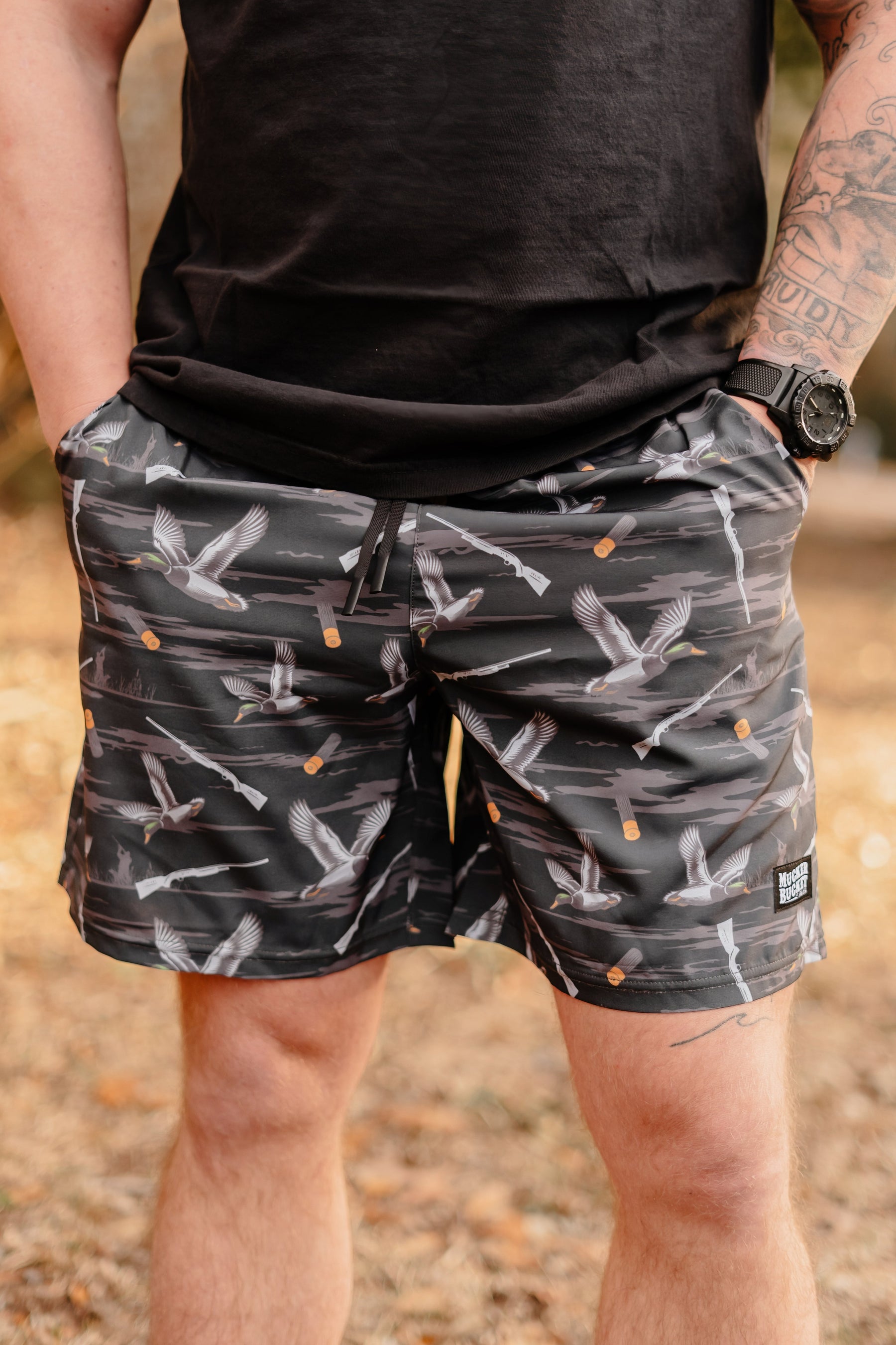 In a natural setting with fallen leaves, a person stands outdoors with hands in pockets, wearing Mucker Buckets black Duck, Duck Boom shorts and a wristwatch. They sport a tattoo on their left arm and are dressed in a black shirt.