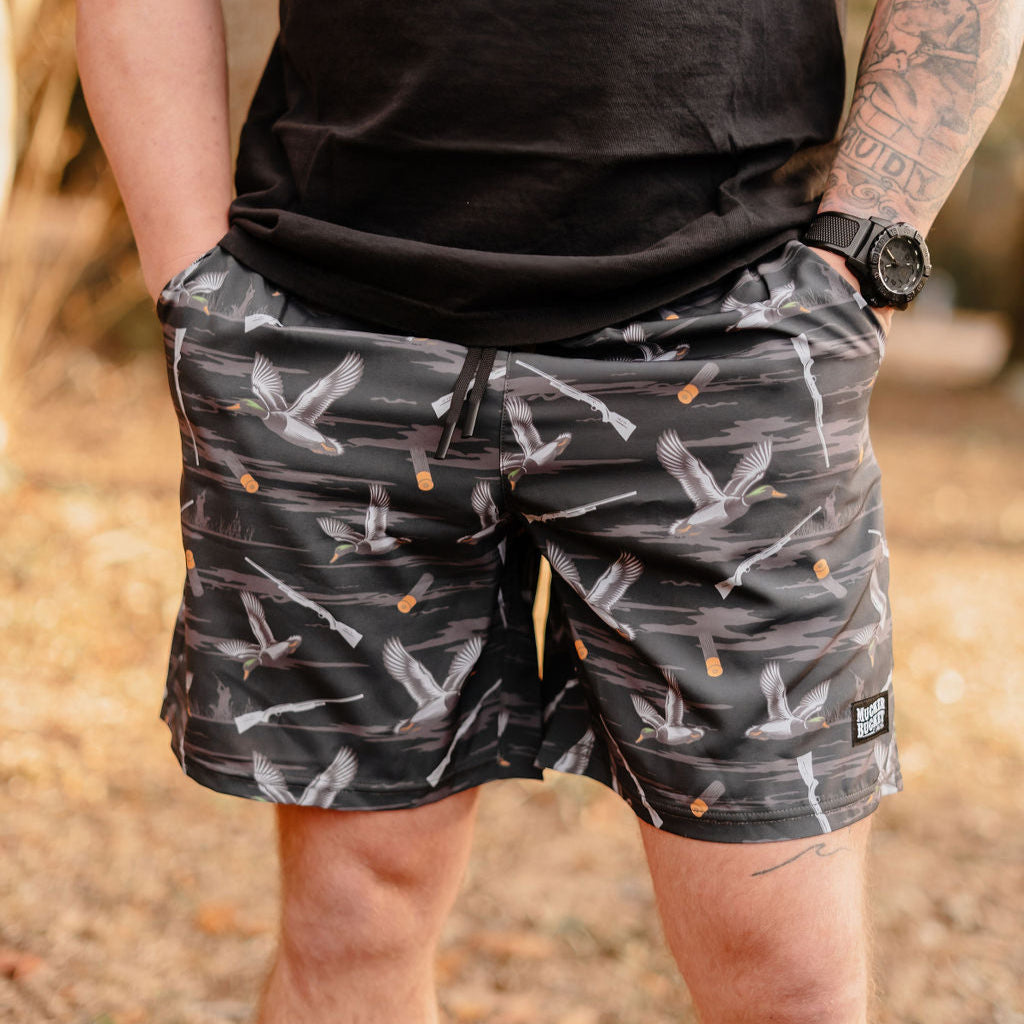 A person outdoors in Mucker Buckets Mens Performance Athletic Shorts, 5.5- Duck, Duck Boom, with a crane pattern and dark drawstring, wears a black watch and T-shirt. The earthy-toned background is blurred.