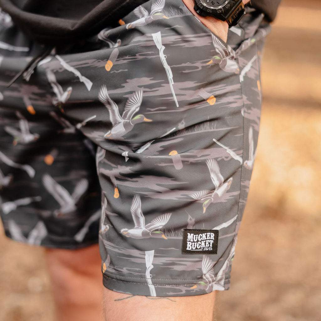 A person outdoors is wearing Mucker Buckets Mens Performance Athletic Shorts in the Duck, Duck Boom pattern, featuring gray camouflage with ducks and shotguns. Part of their hand is visible.