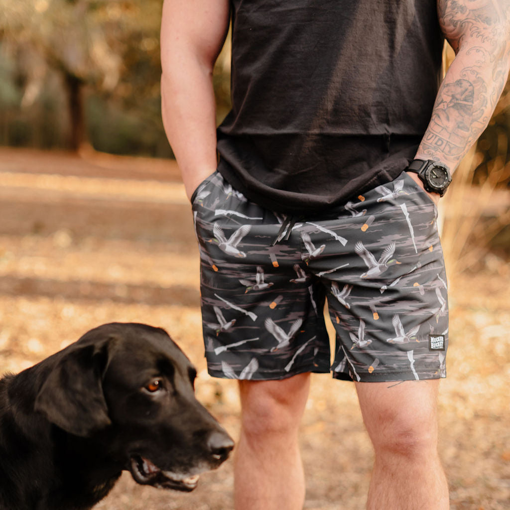 A person in a black shirt and Mucker Buckets Mens Performance Athletic Shorts | 5.5- Duck, Duck Boom stands outdoors with hands in pockets. A black dog is beside them, against a backdrop of trees and soft sunlight.