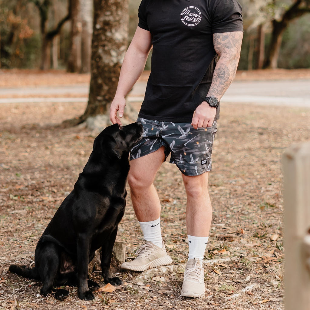A man in a black t-shirt and Mucker Buckets Mens Performance Athletic Shorts | 5.5- Duck, Duck Boom stands outdoors petting a black Labrador beside him, surrounded by trees and scattered dry leaves.