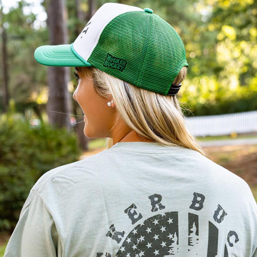 A person with long blonde hair is seen from behind, wearing a green and white Duck It Green Trucker Hat by Mucker Bucket and a light green shirt featuring a high-quality screen-printed circular logo on the back. Trees and a white fence blur in the background.
