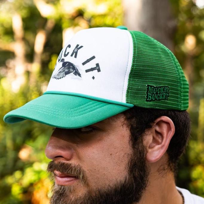 A man wearing the Duck It Green Trucker Hat, by Mucker Bucket, with a mesh back and BUCK IT text alongside a flying bird graphic on the front. The side features a green MUCKER BUCKET logo. This standout hat contrasts against blurred green foliage.