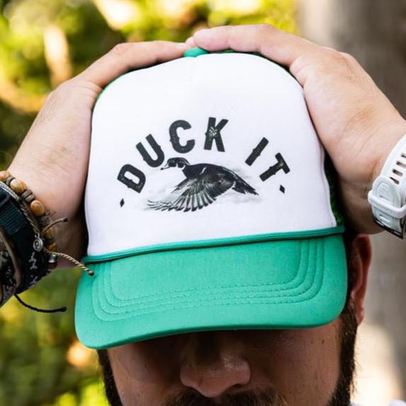 A person wears the Duck It Green Trucker Hat by Mucker Bucket, featuring a white and green design with DUCK IT and a flying duck in high-quality screen print. Their hands, decorated with bracelets and a watch, rest atop the cap against a backdrop of blurred greenery.
