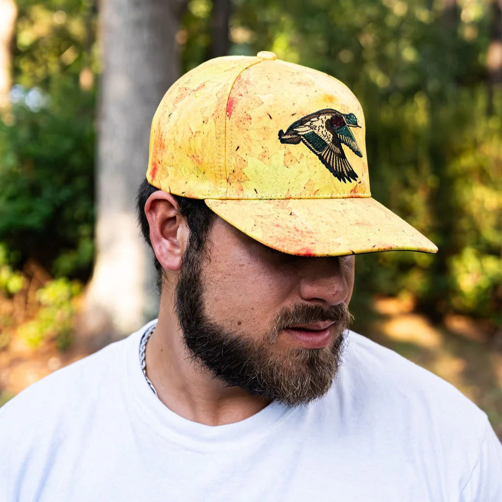 A bearded man in a white t-shirt and yellow Mucker Bucket Golden Leaf Camo Duck Bucket hat with a bird design looks down, set against a blurred outdoor scene of trees and greenery, suggesting serene duck hunting adventures.